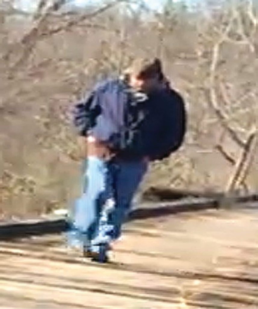 A stabilized, enhanced image of a vaguely male figure wearing a blue coat and jeans walking on the railroad ties of a bridge.