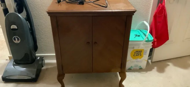 A brown, wood Singer 48 cabinet closed up, between a vacuum and a box of cat litter.